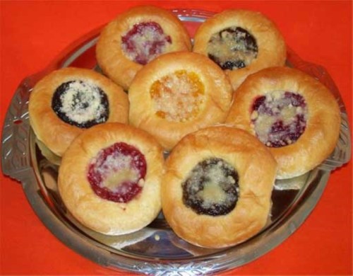 A silver platter holds eight round pastries with various fruit fillings, including red, black, and orange, on an orange background.