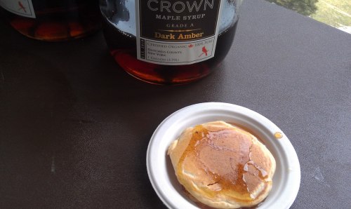 A small pancake drizzled with dark amber maple syrup on a white plate, with a bottle of syrup in the background.