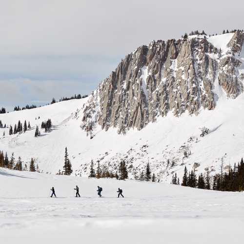 Four skiers traverse a snowy landscape with a rocky mountain backdrop under a cloudy sky.