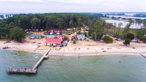 Aerial view of a beach with a pier, surrounded by trees and parked cars, with people enjoying the water and sand.