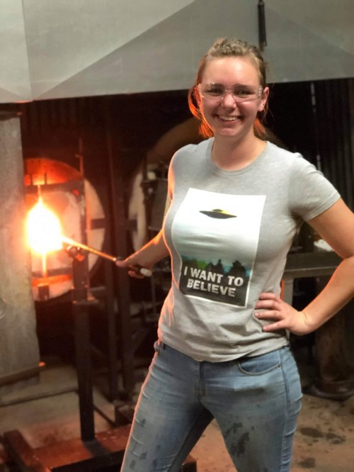A smiling woman in a "I Want to Believe" shirt stands near a glowing furnace, holding a tool in a glassblowing studio.