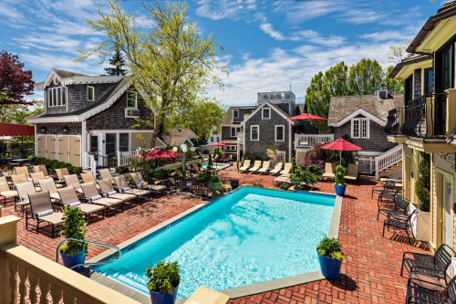 A sunny pool area with lounge chairs, surrounded by greenery and charming buildings, featuring red umbrellas and blue planters.