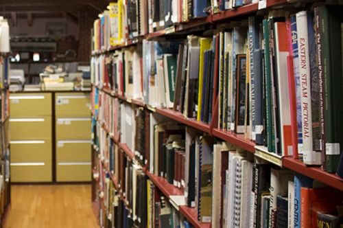 A library aisle lined with bookshelves and filing cabinets in the background.
