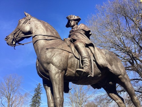 A bronze statue of a mounted figure in historical attire against a clear blue sky, surrounded by trees.