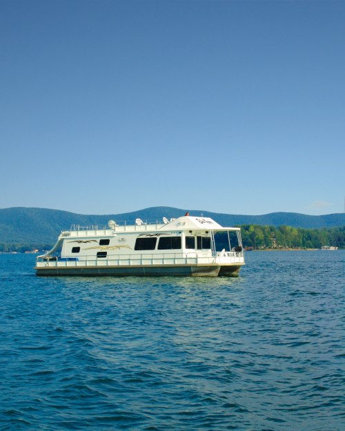 A houseboat floats on calm water with mountains in the background under a clear blue sky.