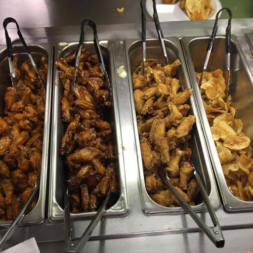 A buffet display of various chicken wings in metal trays, with tongs, alongside a tray of potato chips.