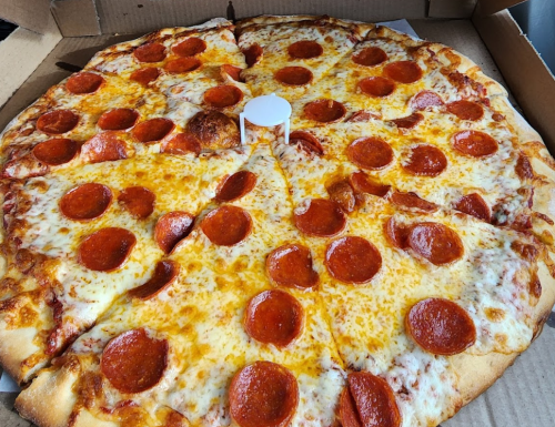 A large pepperoni pizza with melted cheese, sliced into wedges, in a pizza box with a white pizza saver in the center.
