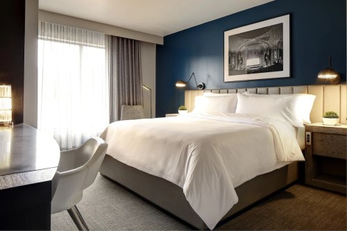 A modern hotel room featuring a king-sized bed, desk, and large window with sheer curtains.