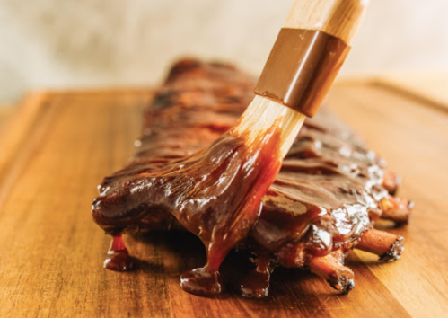 A close-up of a glazed rib being brushed with barbecue sauce on a wooden cutting board.