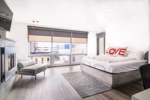 A modern hotel room with a large bed, decorative "LOVE" pillows, a cozy chair, and a view through large windows.