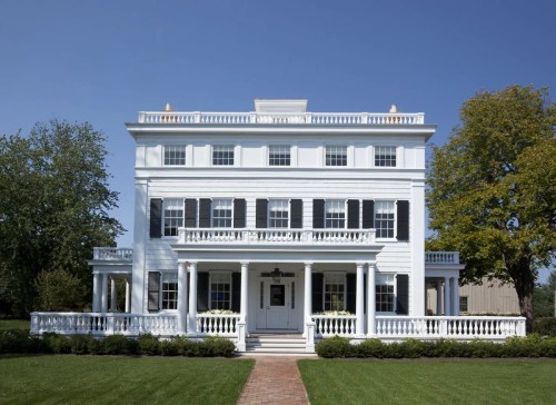 A large white historic house with a porch, green lawn, and blue sky in the background.