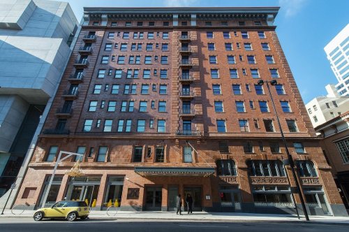 Historic brick building with large windows, featuring a modern structure beside it and a yellow car parked in front.