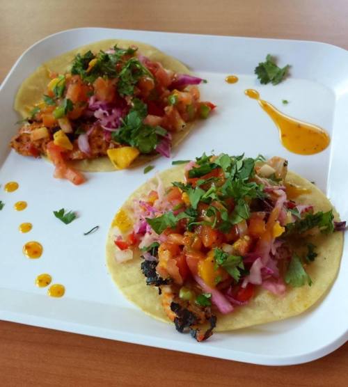 Two tacos on a white plate, topped with fresh salsa, onions, cilantro, and drizzled with sauce.