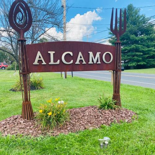 Sign for Alcamo with decorative elements, surrounded by flowers and grass, under a blue sky with clouds.