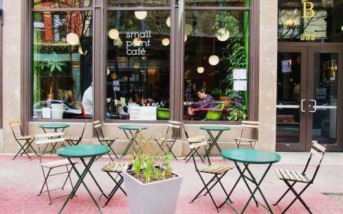 A cozy café with green tables and chairs outside, large windows showcasing a lively interior and plants.