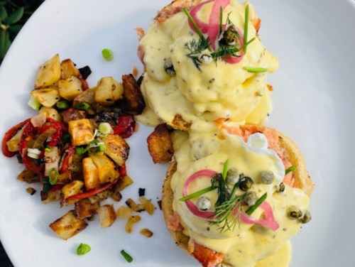 A plate of eggs Benedict topped with hollandaise sauce, capers, and pickled onions, served with a side of sautéed potatoes.