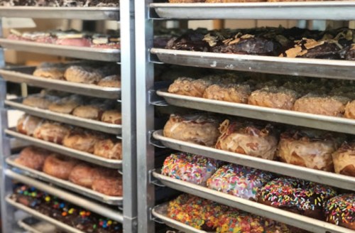 A display of various donuts on metal trays, featuring colorful sprinkles, chocolate glaze, and different toppings.