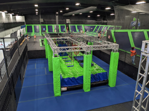 A vibrant indoor trampoline park featuring green and blue obstacle courses and trampolines, with people enjoying the space.