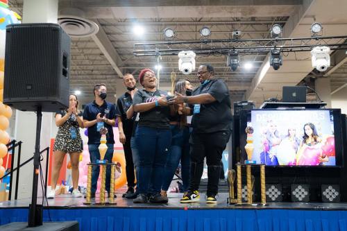 A group celebrates on stage with trophies, smiling and applauding at an event, with colorful decorations in the background.