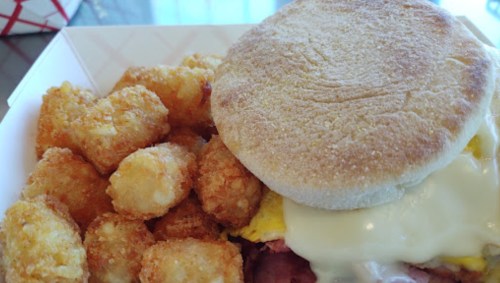 A breakfast sandwich with melted cheese on a toasted English muffin, served with crispy tater tots on the side.
