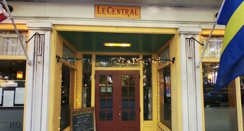 Entrance of Le Central restaurant, featuring yellow trim, a welcoming door, and decorative lights.