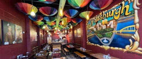 Colorful umbrellas hang from the ceiling of a cozy restaurant, featuring a mural of Pittsburgh and wooden tables.