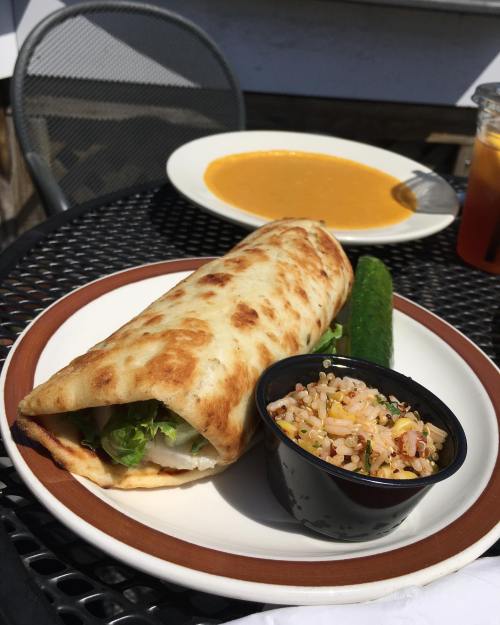 A plate featuring a wrapped flatbread with lettuce, a side of cucumber, and a small bowl of rice salad, with soup in the background.