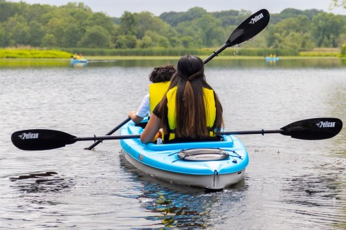 A person paddles a blue kayak with a child, both wearing yellow life jackets, on a calm lake surrounded by greenery.