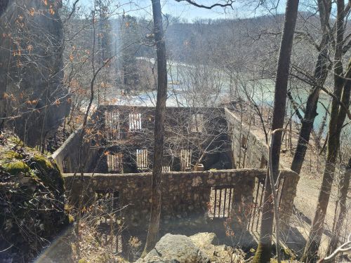 Ruins of a stone building surrounded by bare trees, overlooking a river in a wooded area.