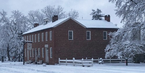 A historic brick building covered in snow, surrounded by snow-laden trees and a quiet, wintry landscape.