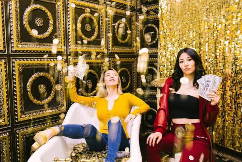 Two women in a glamorous setting, one sitting in a bathtub with money, the other holding cash, surrounded by gold decor.