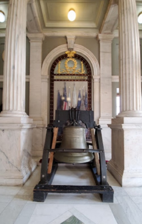 A large bell displayed in a marble hall, framed by columns and flags, with a decorative backdrop.