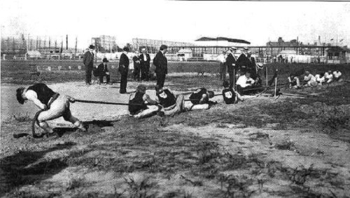 Historical photo of athletes competing in a tug-of-war event, with spectators observing in the background.