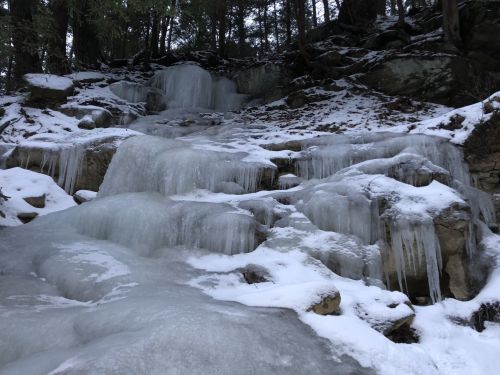 Frozen waterfall cascading over rocky terrain, surrounded by snow and trees in a winter landscape.