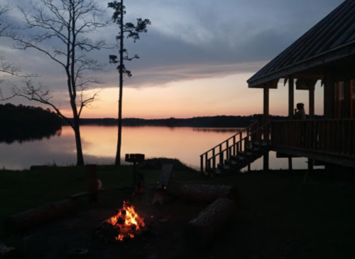 A serene lakeside scene at sunset, featuring a campfire and a cabin with a porch overlooking the water.