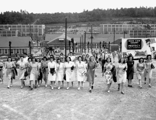 A group of women in dresses walking together, with buildings and power lines in the background. Black and white photo.