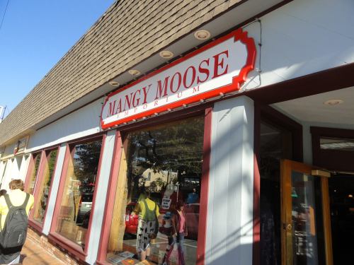 Sign for "Mangy Moose Emporium" on a storefront, with large windows and a clear blue sky above.