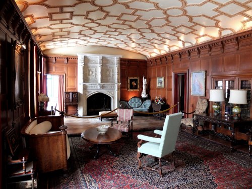 A spacious, elegantly furnished room with wooden walls, a decorative ceiling, and a fireplace, featuring vintage furniture.
