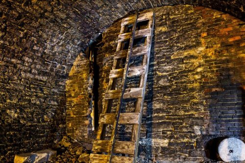A wooden ladder leans against a stone wall in a dimly lit, arched underground space.