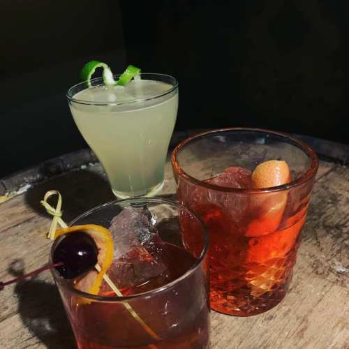 Three colorful cocktails on a wooden surface: a green drink with a lime twist, and two amber drinks with garnishes.
