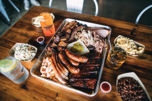 A large platter of assorted barbecue meats, sides, and drinks on a wooden table.