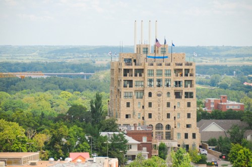 A tall, stone building with flags on top, surrounded by greenery and distant hills under a blue sky.
