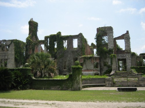 Ruins of a stone building overgrown with vines and greenery, set against a clear blue sky.