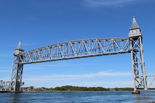 A silver lift bridge with towers against a clear blue sky, spanning over calm water with greenery in the background.
