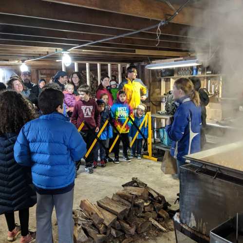 A group of children and adults watch a demonstration in a rustic setting, with steam rising from a large pot.