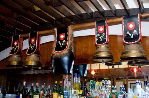 Five decorative bells hang from a wooden bar, adorned with Swiss flags and floral designs, above a colorful array of bottles.