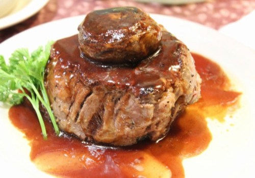 A plated beef dish topped with a rich sauce and a small round garnish, served with a sprig of parsley.