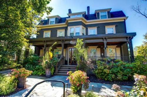 A charming, multi-story house with a porch, surrounded by lush greenery and colorful flowers.