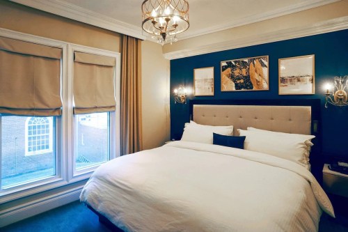 Cozy bedroom with a large bed, elegant decor, and two windows with shades, featuring artwork on the wall.