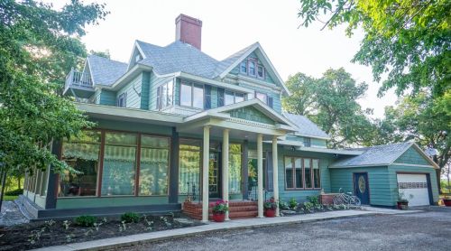 A large, green Victorian-style house with a porch, surrounded by trees and a driveway.